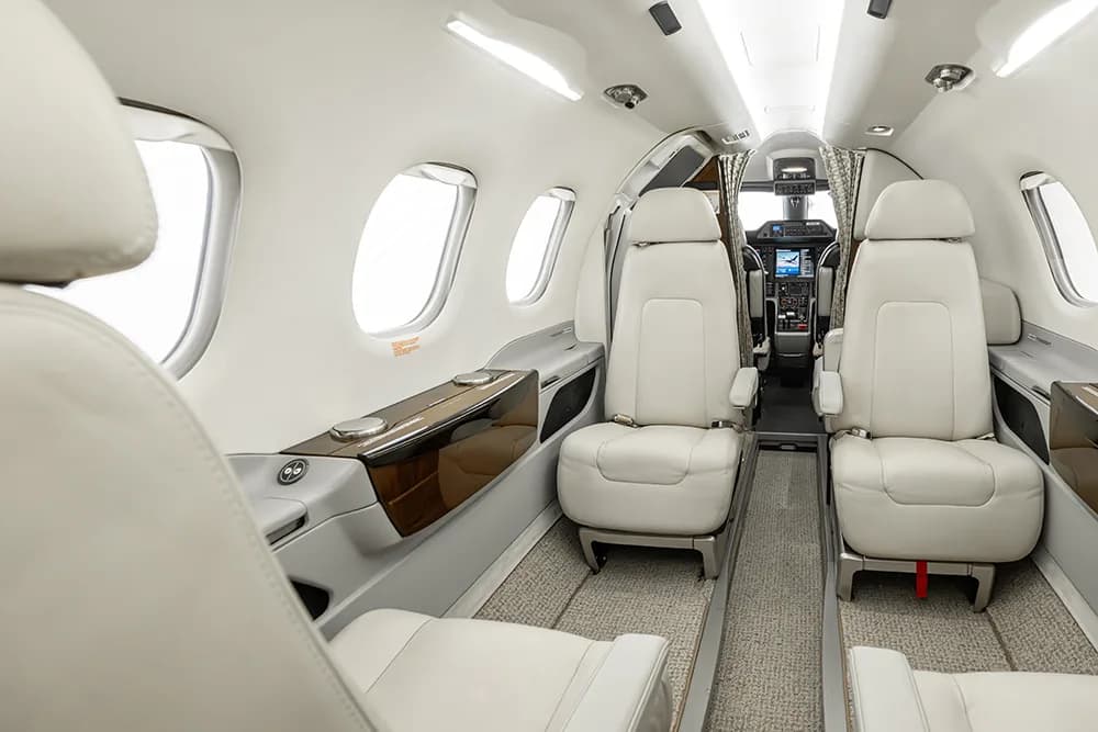 The interior of a private jet cabin with white leather seats and dark wood accents, looking towards the cockpit.
