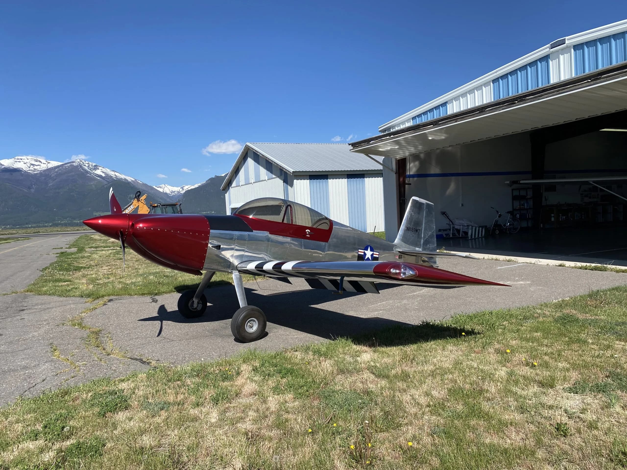A shiny silver and red Vans RV-8 aircraft with black and white invasion stripes on the wings, parked outside a hangar with mountains in the background.