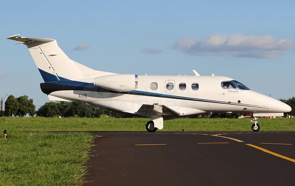 A white Embraer Phenom 300 business jet with a blue and black stripe is seen from the side on an airfield.