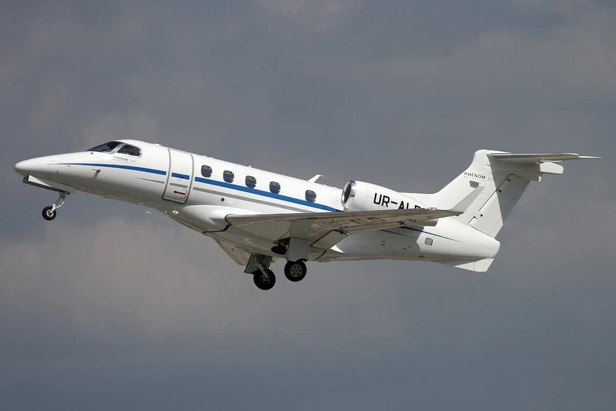 A white Embraer Phenom 300 is shown in flight with landing gear down.