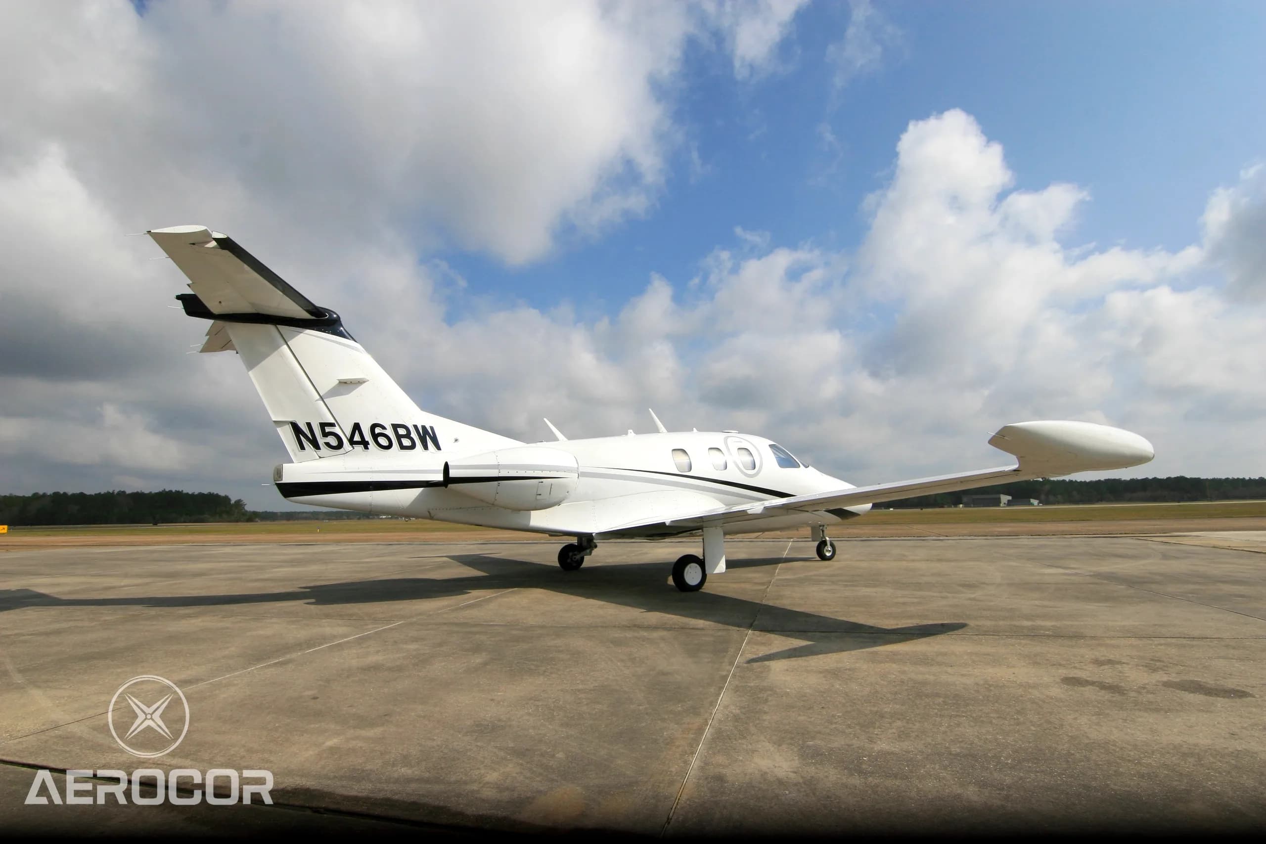 A white Eclipse 500 jet with black and grey stripes parked on an airfield under a cloudy blue sky.
