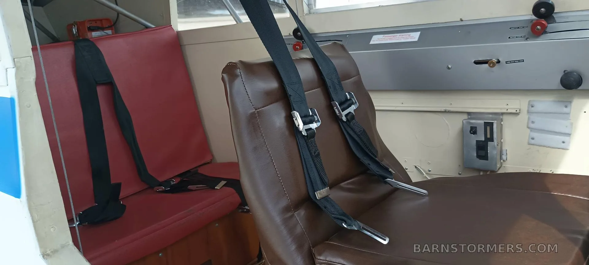 The interior of an aircraft cabin showing a brown leather pilot seat and a red passenger seat with seatbelts.