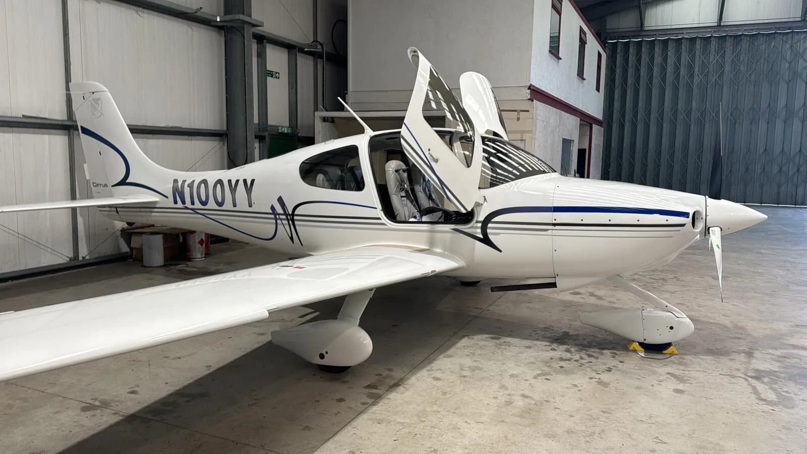 A white Cirrus SR22 aircraft with blue and grey stripes parked inside a hangar with its gull-wing doors open.