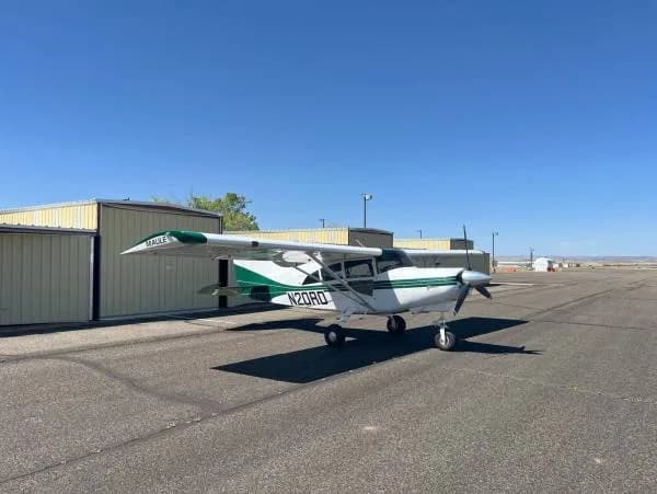 A white and green Maule aircraft, registration N20RD, parked on a tarmac next to hangars.