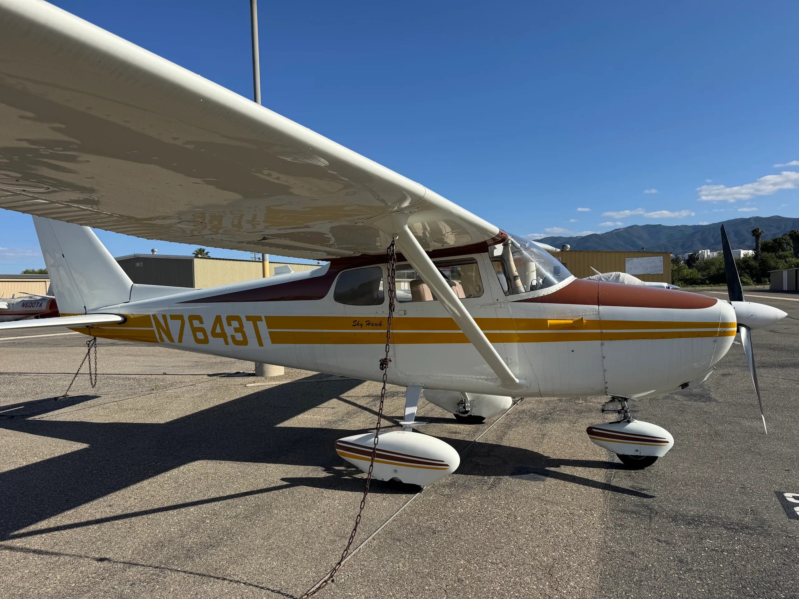 A white and brown Cessna Skyhawk with yellow stripes, registration N7643T, parked on an asphalt tarmac under a clear blue sky.