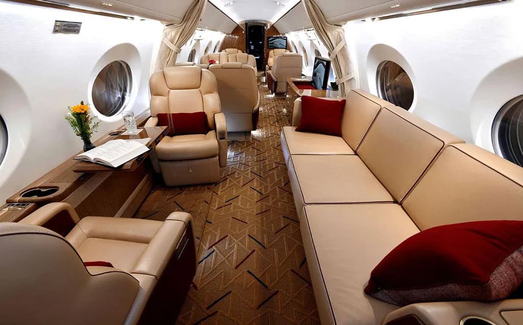 A wide view of a private jet's luxurious cabin interior with a beige sofa, individual seats, and red accent pillows.