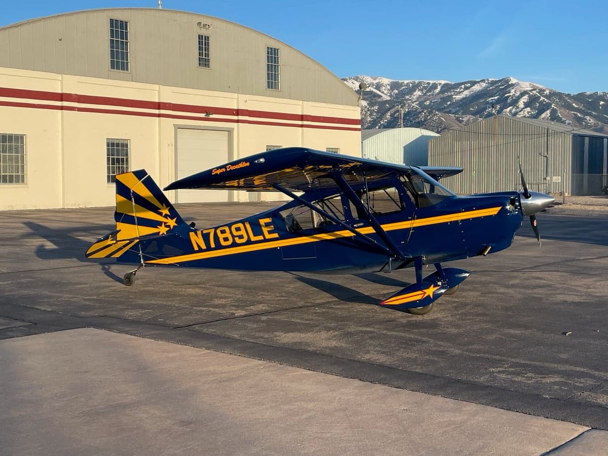 A blue and yellow Super Decathlon aircraft with registration N789LE parked on an airfield with hangars and snowy mountains in the background.