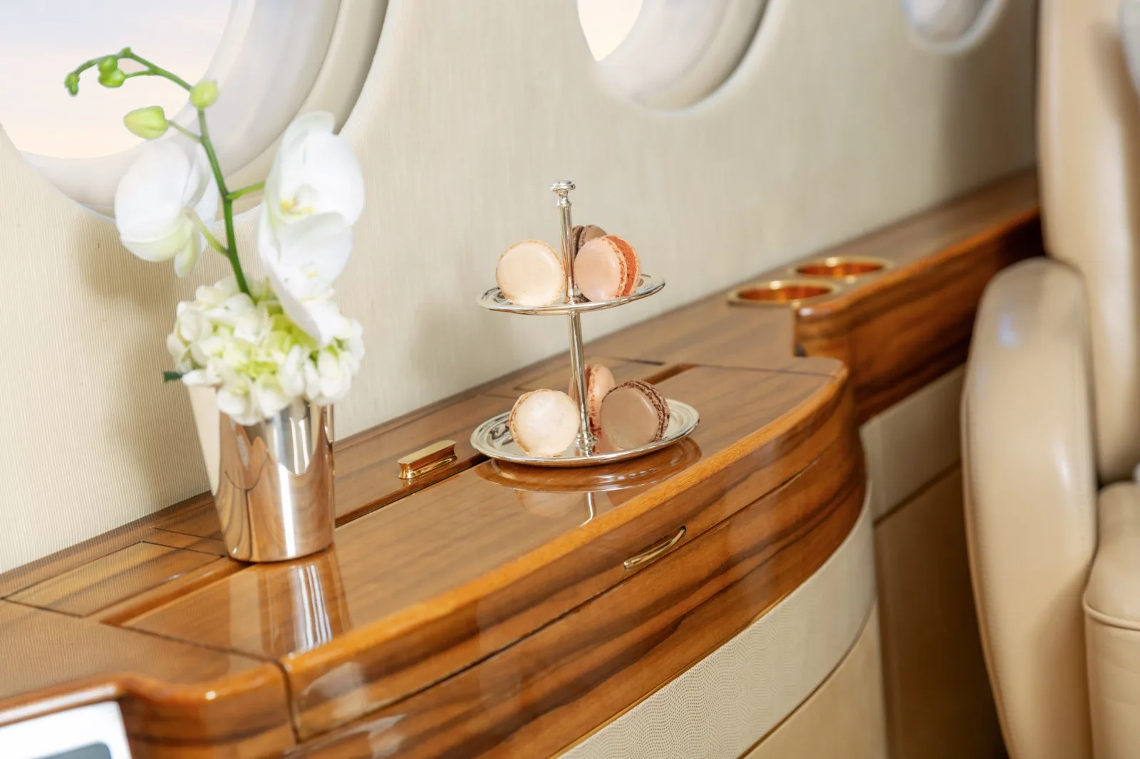 A close-up of a wooden side table in the aircraft cabin, featuring a vase of white orchids and a two-tier stand with macarons.