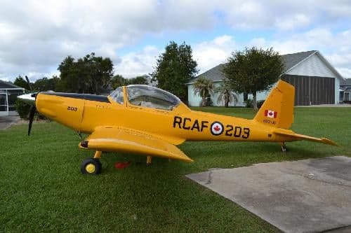 A yellow aircraft with RCAF markings is parked on a grassy lawn in front of a house.