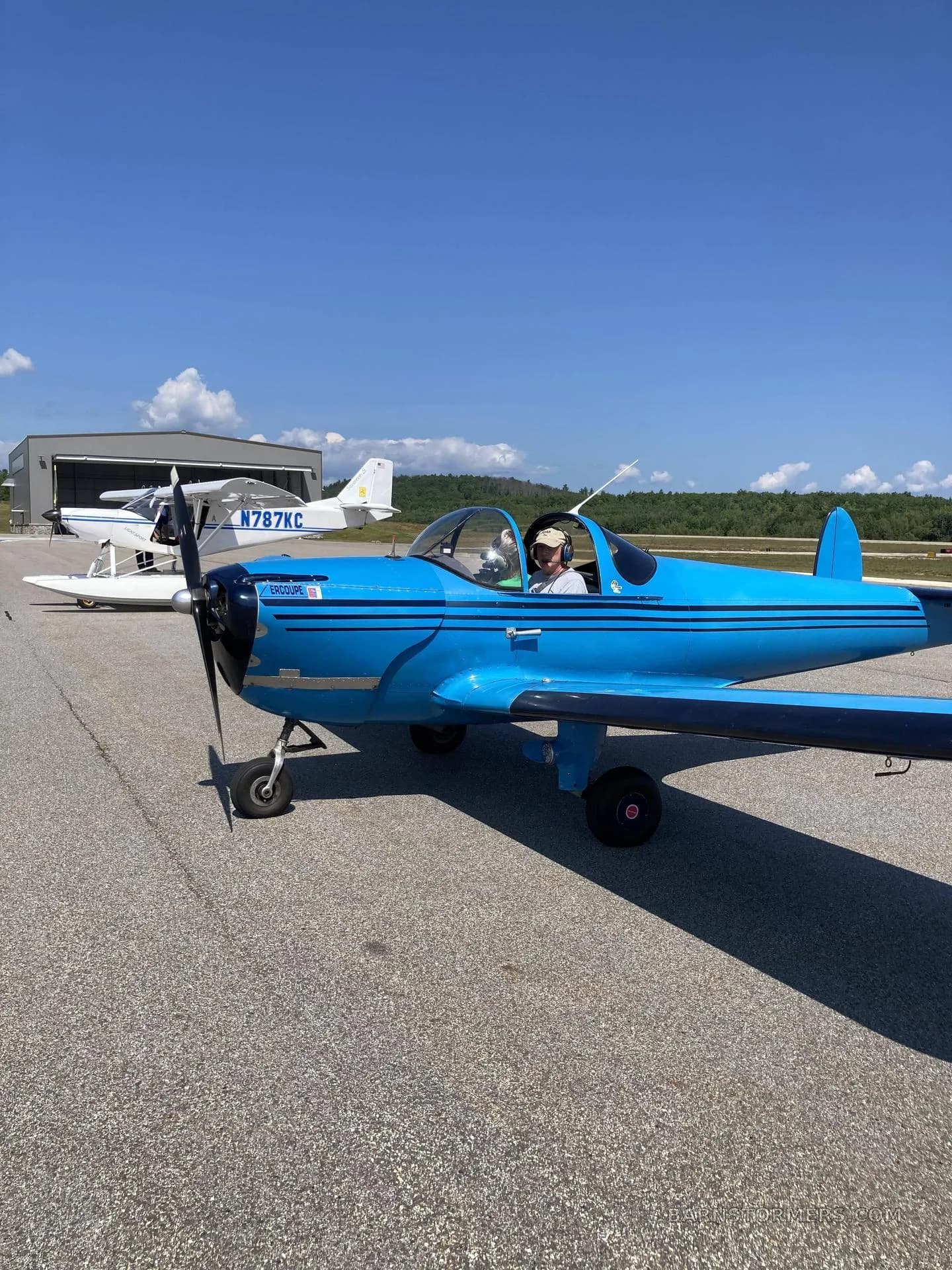 A bright blue Ercoupe aircraft with black stripes and a pilot in the cockpit is parked on an airfield with other planes and hangars in the background.