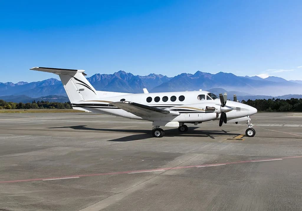 A white twin-turboprop aircraft with black and gold stripes is parked on a tarmac with mountains in the background.