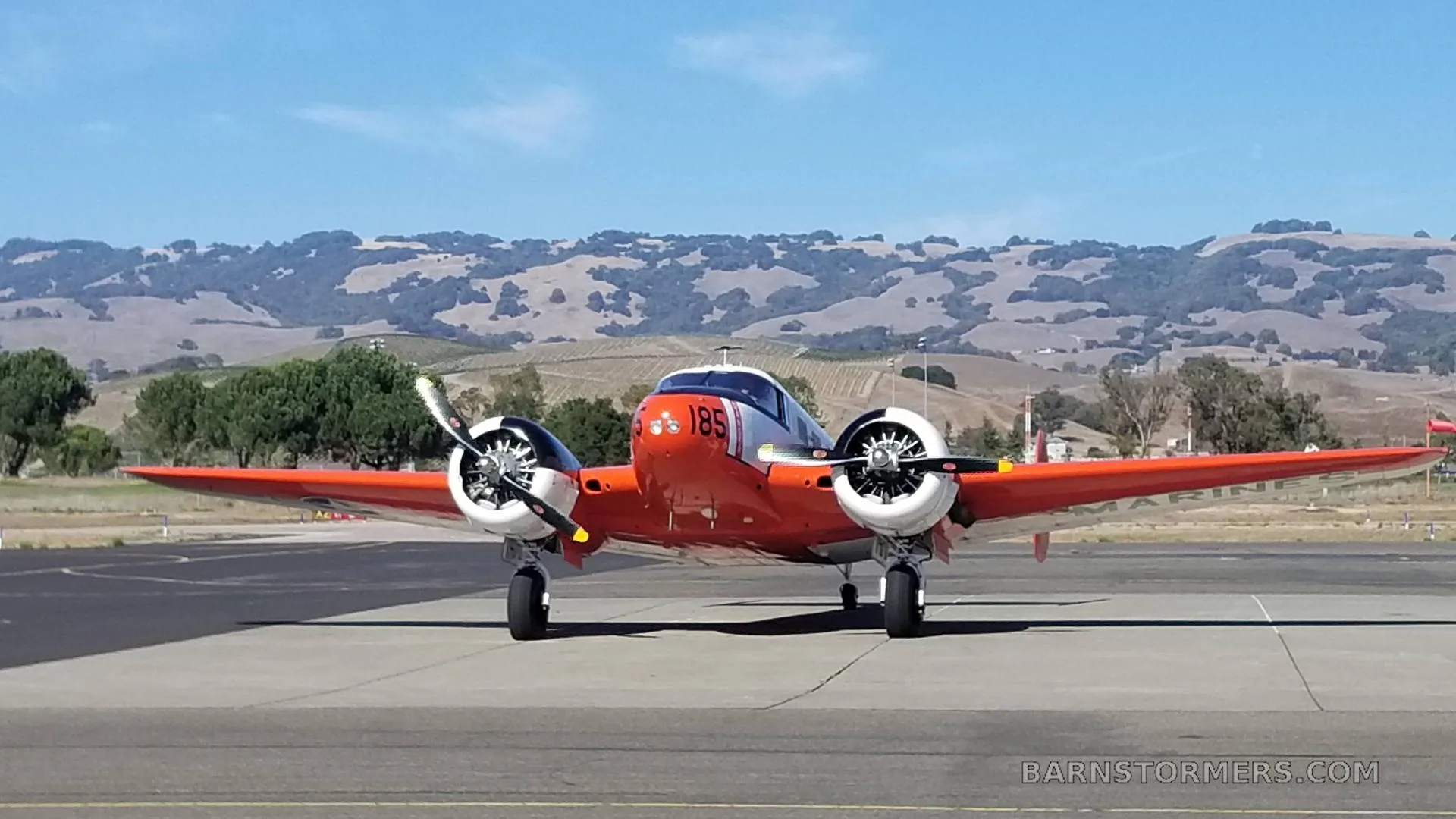 A front view of an orange and white twin-engine aircraft with '185' on the nose, parked on an airfield with hills in the background.