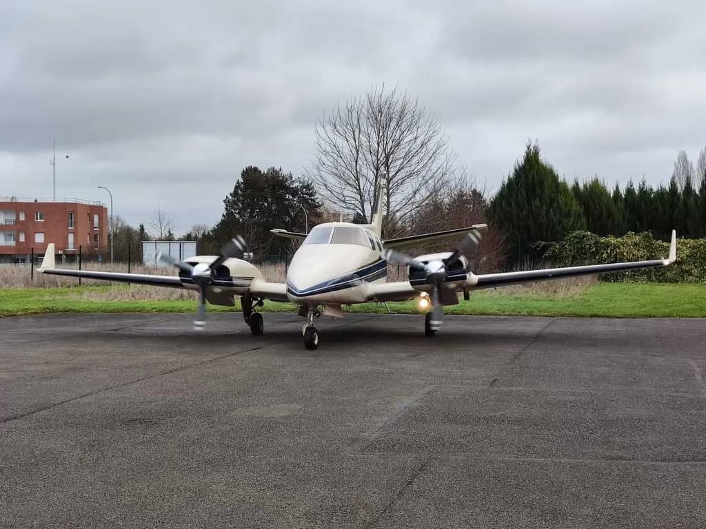 A beige and blue twin-engine Piper PA-31-350 Navajo Chieftain aircraft is parked on an asphalt surface with trees and buildings in the background.
