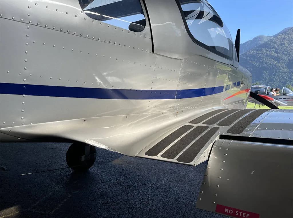 Close-up of the silver fuselage of a Sonaca 200 aircraft showing the blue stripe, step pads, and a "NO STEP" warning.