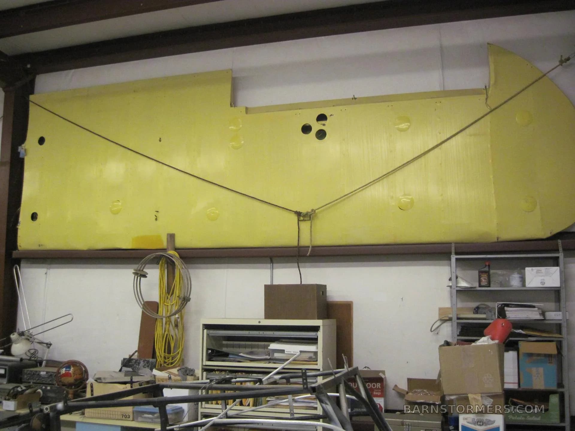 A large yellow aircraft wing with cables attached, stored on a high shelf in a cluttered workshop.