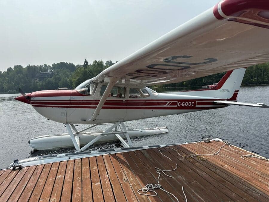 A Cessna floatplane with red and white paint scheme is docked at a wooden pier on a lake.