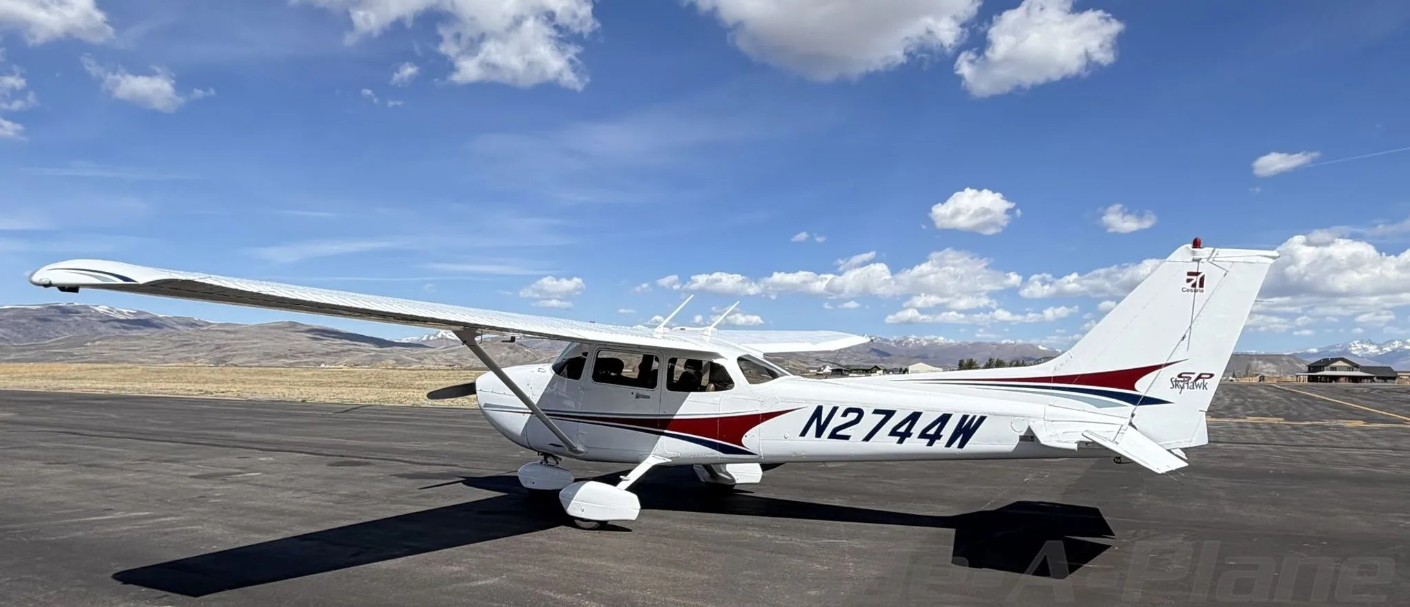 A white Cessna 172 Skyhawk with red and blue stripes, registration N2744W, parked on an airfield under a blue sky.