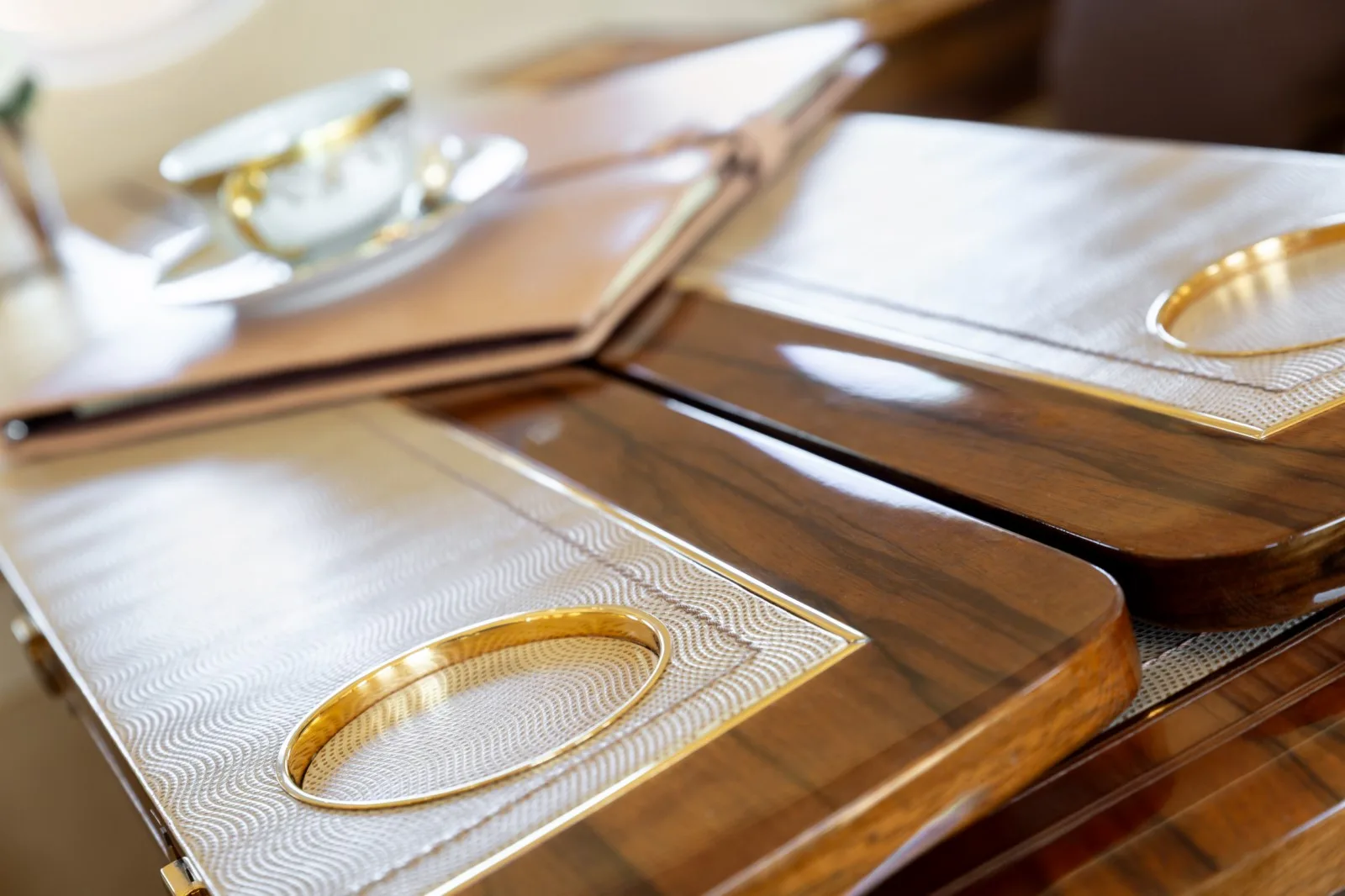 A close-up of a polished wooden table in the aircraft cabin with gold-rimmed placemats and a teacup in the background.