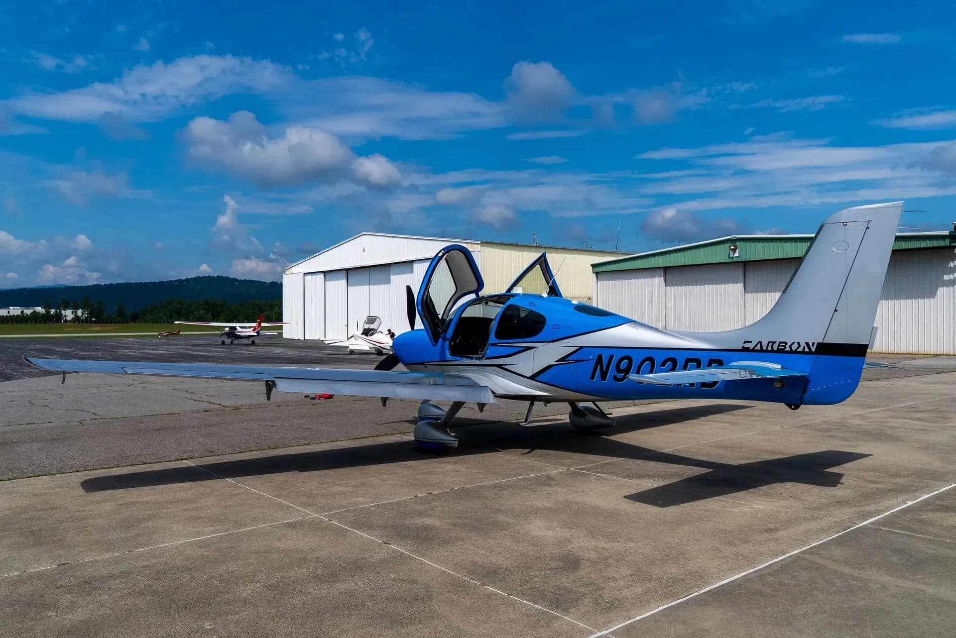A blue and silver Cirrus SR22 aircraft with its doors open, parked on an airfield with hangars and mountains in the background.