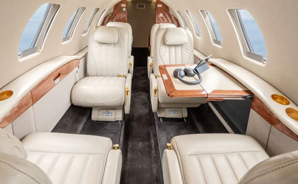 The aircraft's cabin interior features light cream leather seats, a wooden table with a tablet and headphones, and large windows.