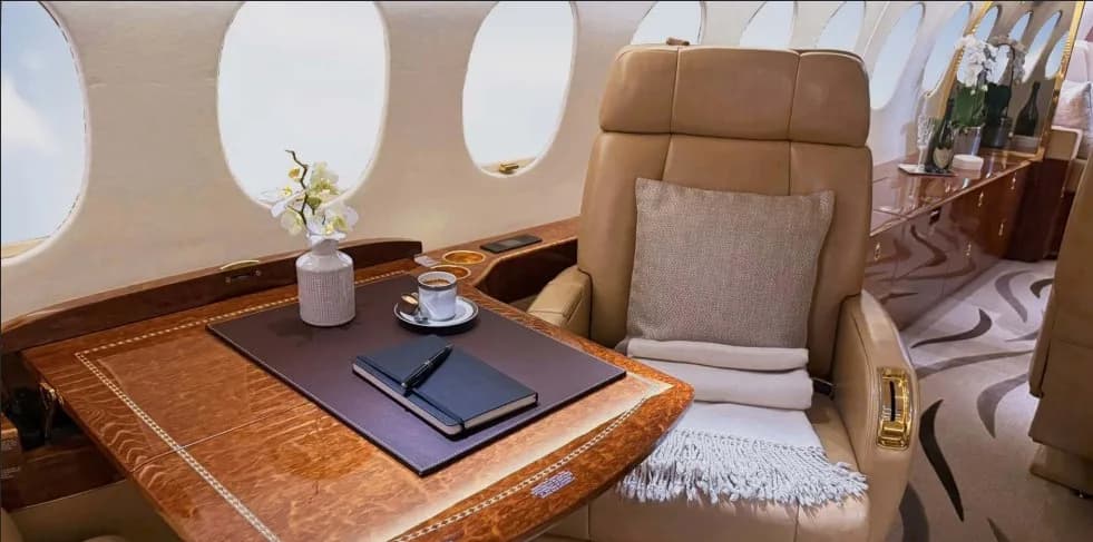 A tan leather armchair next to a polished wooden table with a notebook, pen, coffee cup, and a small vase with flowers in a private jet cabin.