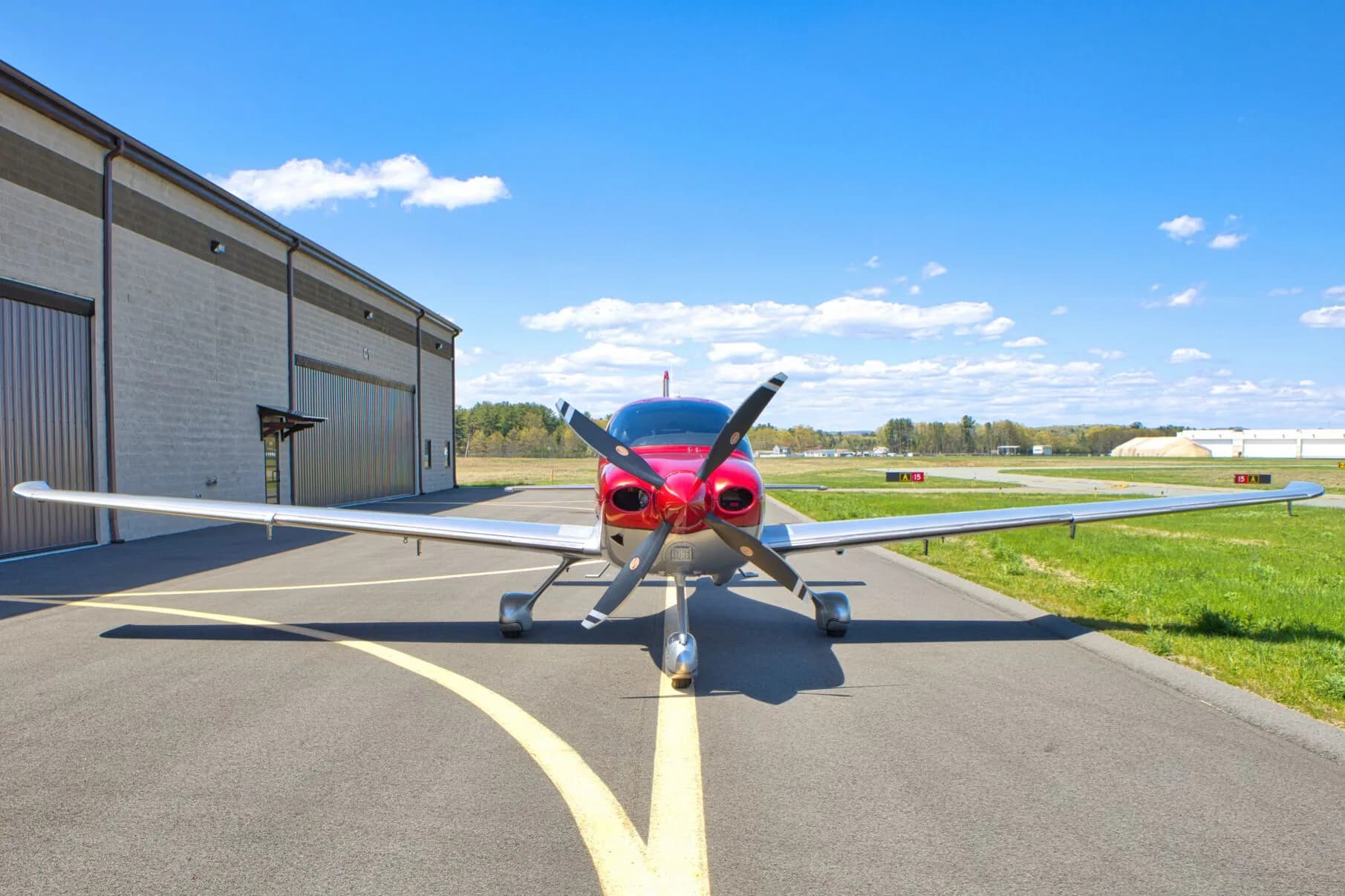 Front view of a red and silver Cirrus SR22T aircraft parked on an airport tarmac with a hangar in the background.