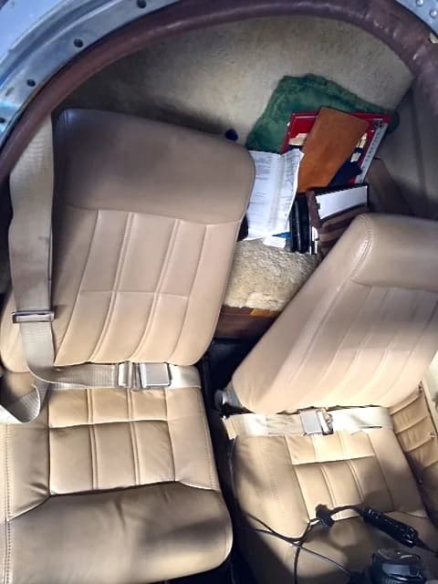 The tan leather seats of a small aircraft cabin, with seatbelts visible and some items stored in the rear.