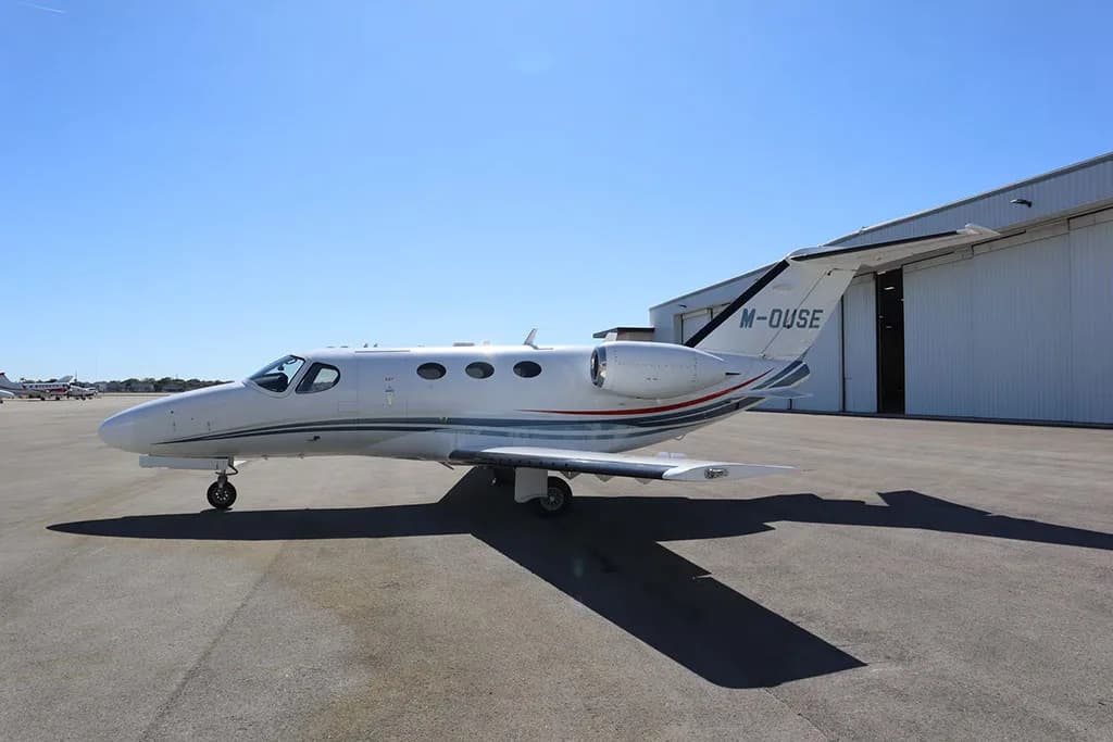 A white business jet with blue and red stripes, seen from a slightly different side angle, parked on an apron under a clear sky.