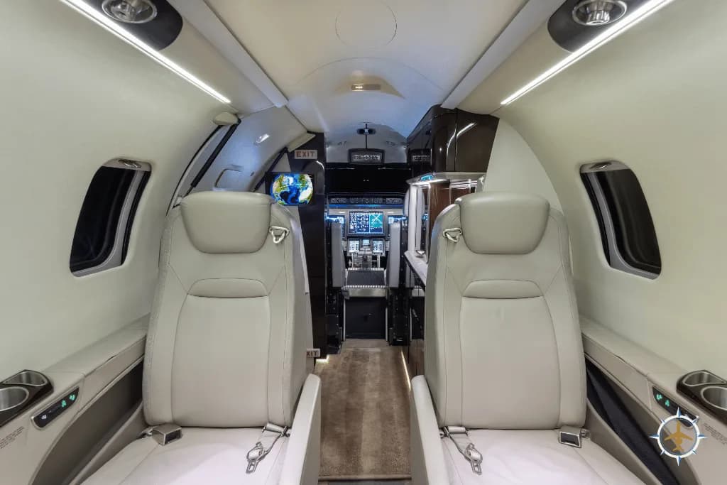 A view from the rear of a private jet cabin, showing light-colored leather seats and the cockpit in the distance.