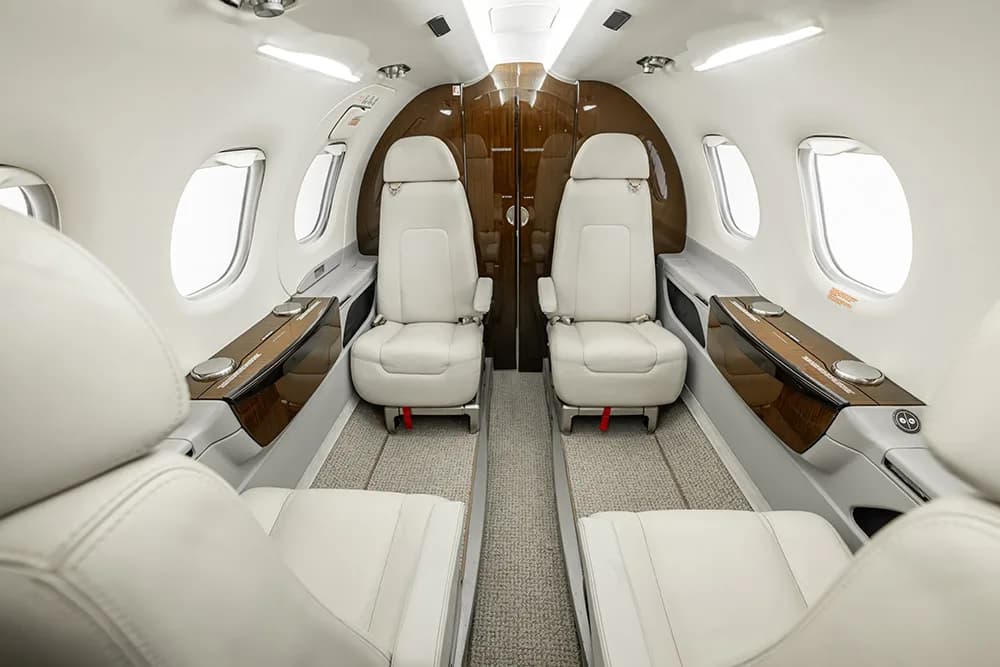 The interior of a private jet cabin with white leather seats and dark wood accents, looking towards the rear.