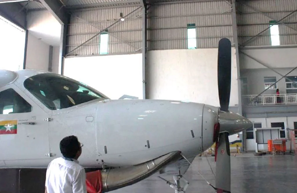 A person stands in front of the propeller and nose of a white aircraft with a Myanmar flag decal, inside a hangar.