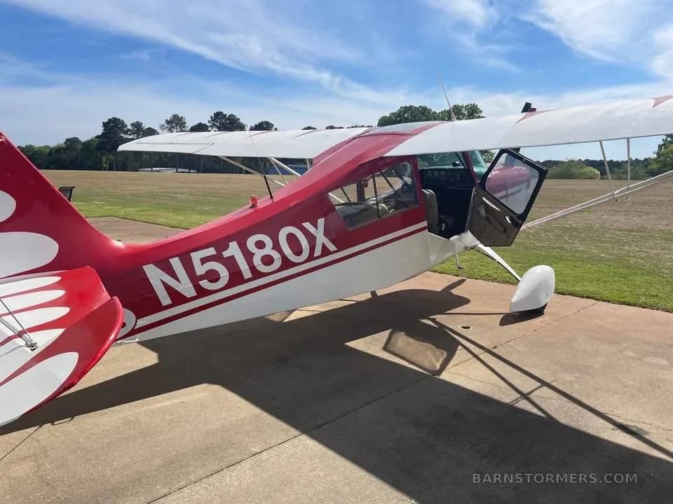 A red and white single-engine aircraft with registration N5180X parked on an airfield with its door open.