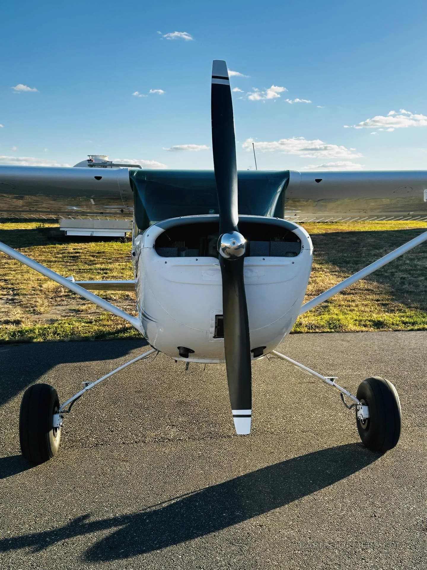 A head-on view of a white Cessna 150/152 type aircraft with a black propeller, parked on an asphalt ramp.
