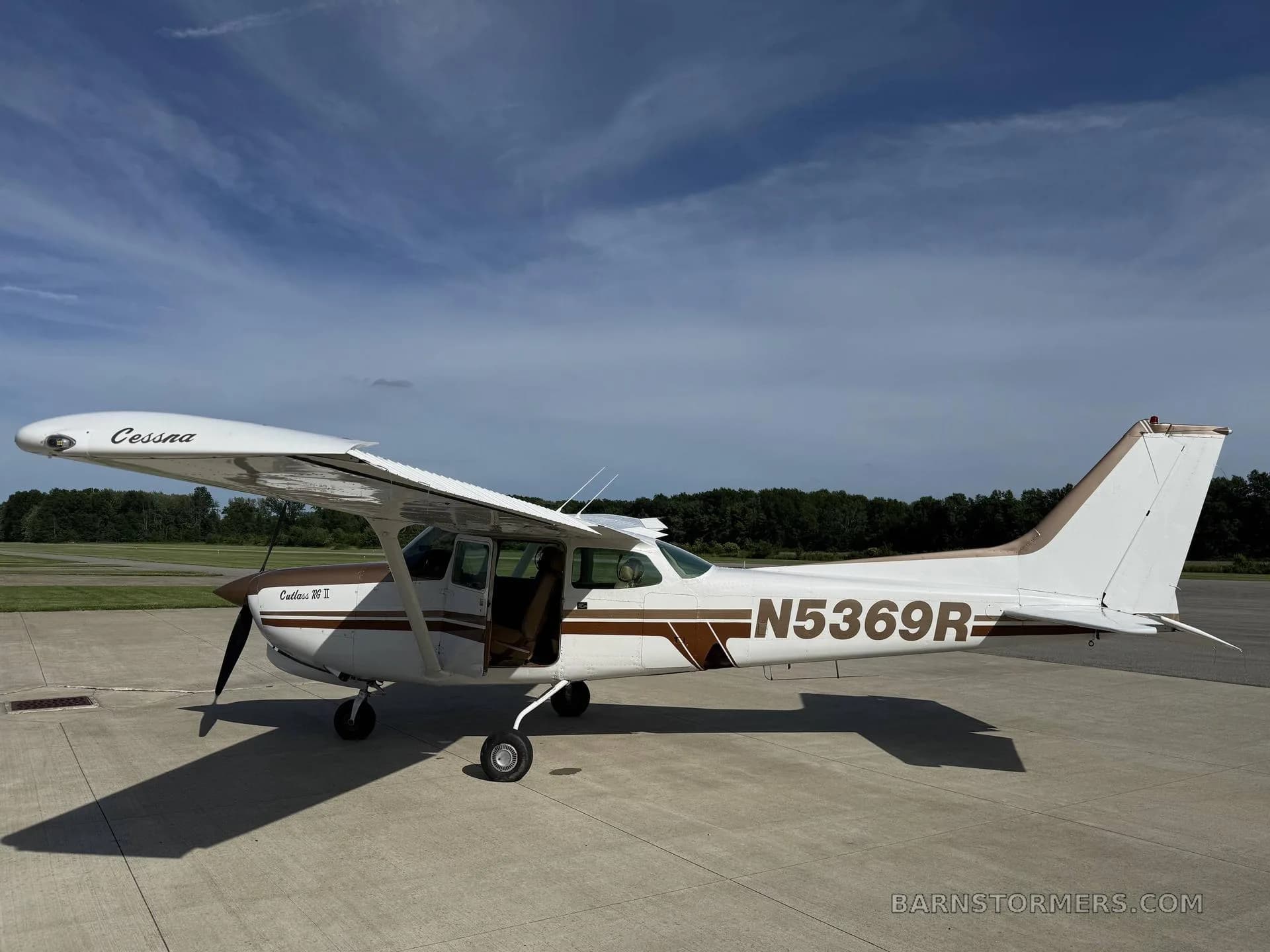 A white and brown Cessna Cutlass RG II (N5369R) is parked on an asphalt ramp with its door open, under a blue sky.