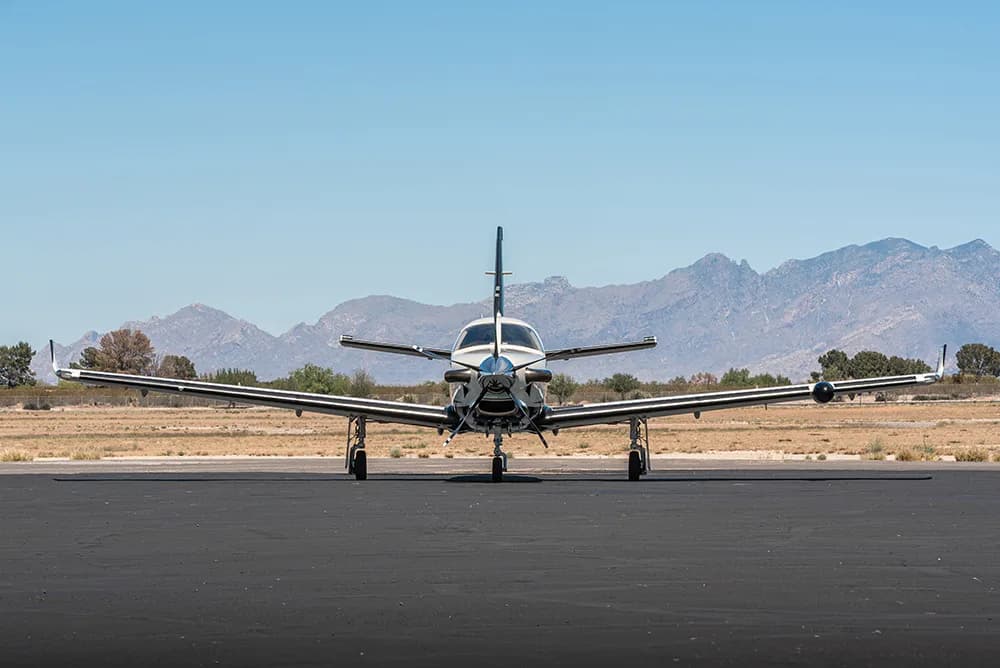 Front view of a white and black TBM 930 aircraft parked on an airfield with mountains in the background.