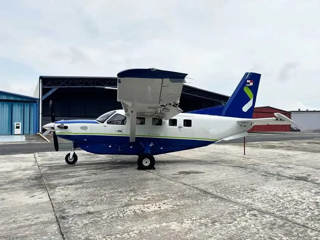 A blue and white Quest Kodiak 100 aircraft with a green stripe and Panamanian flag on the tail, parked on a concrete apron in front of a hangar.