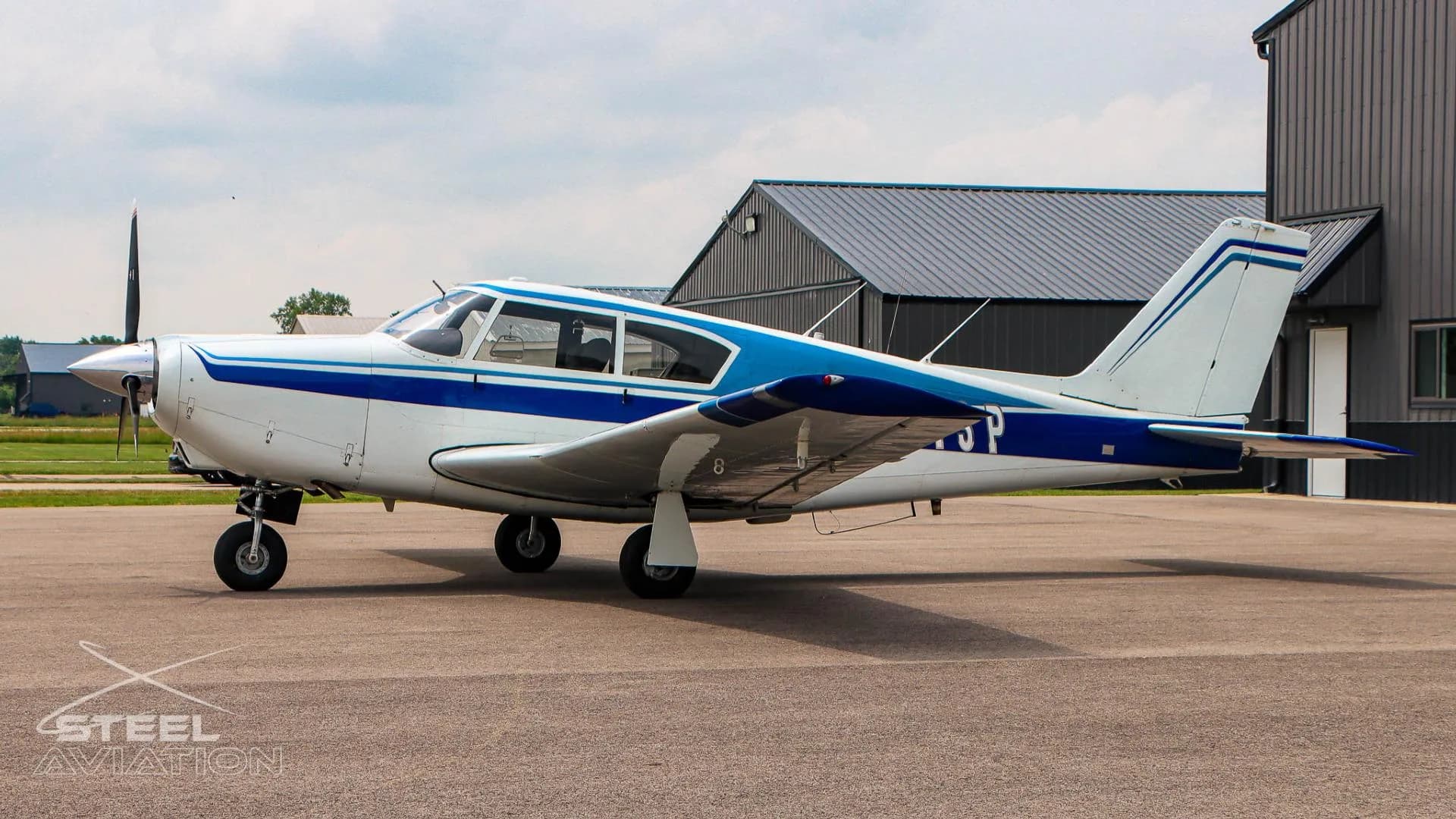 A side view of a white and blue single-engine aircraft with a dark gray hangar in the background.
