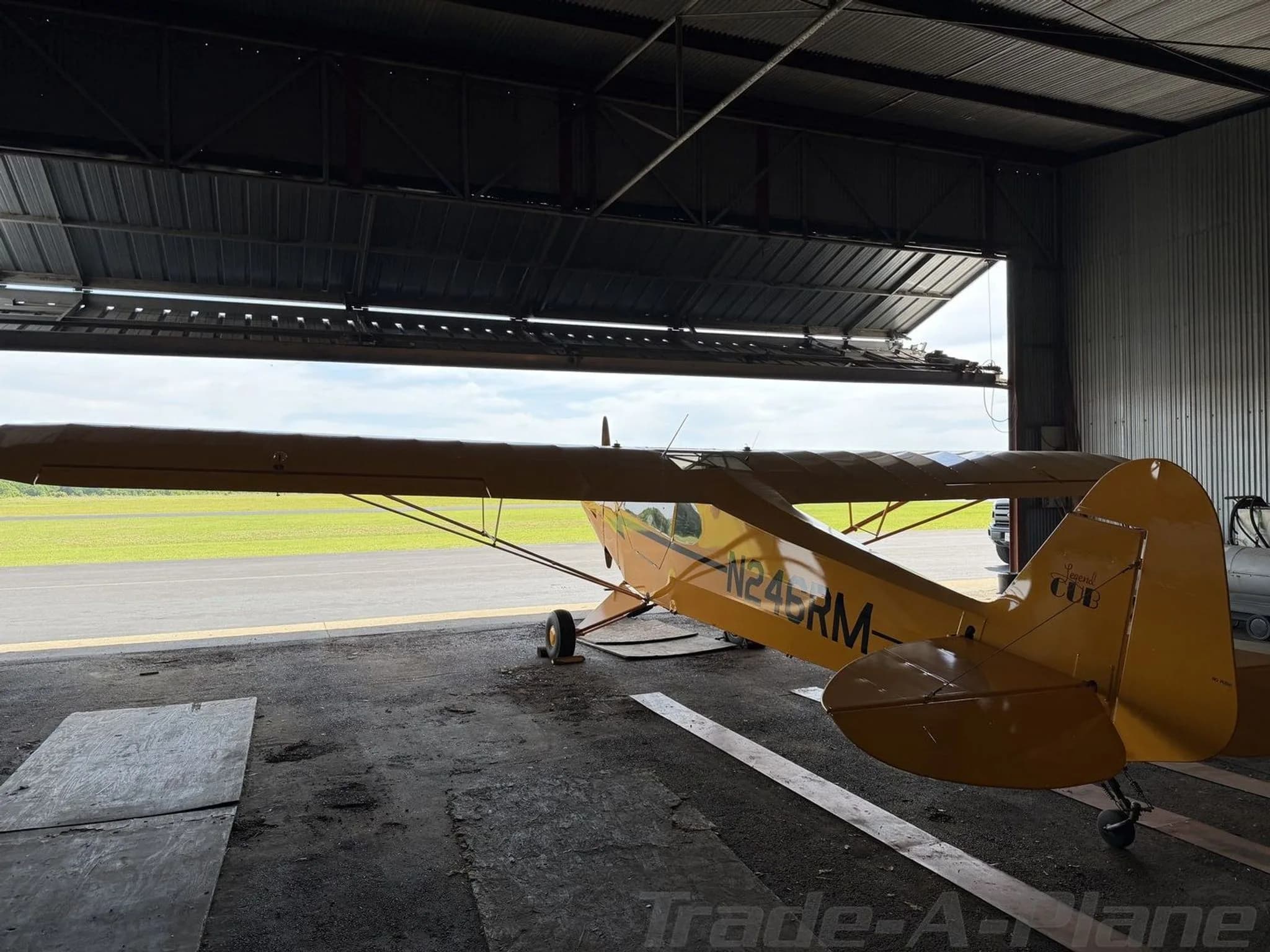 Rear-side view of a yellow American Legend Cub aircraft in a hangar, looking out to a green airfield.