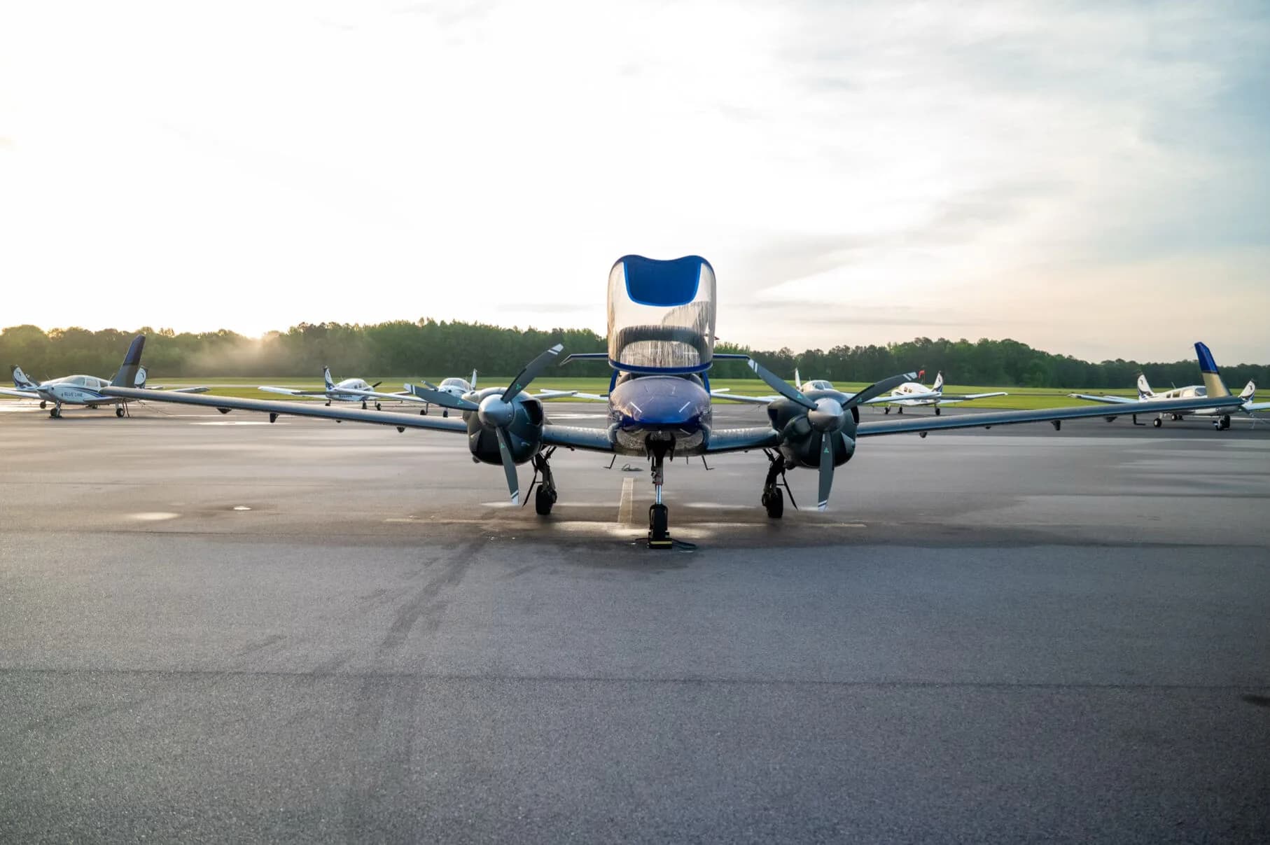 Front view of the blue and grey Diamond DA42NG aircraft with its canopy open, parked on an airfield at sunrise.