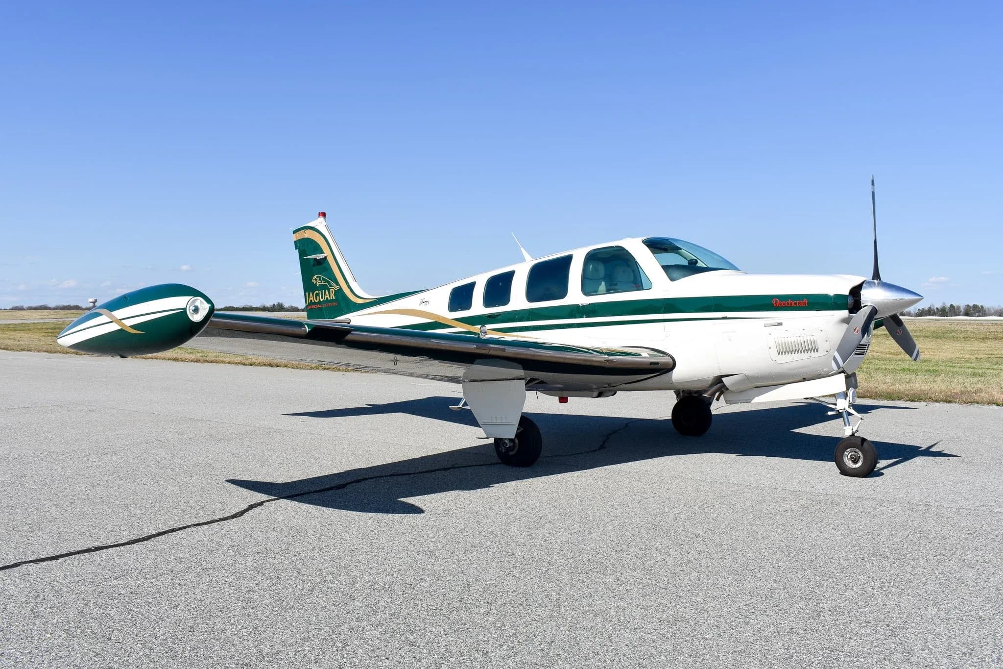 A white and green Beechcraft Bonanza A36 with gold stripes and a "Jaguar Special Edition" logo on the tail, parked on an asphalt ramp under a clear blue sky.