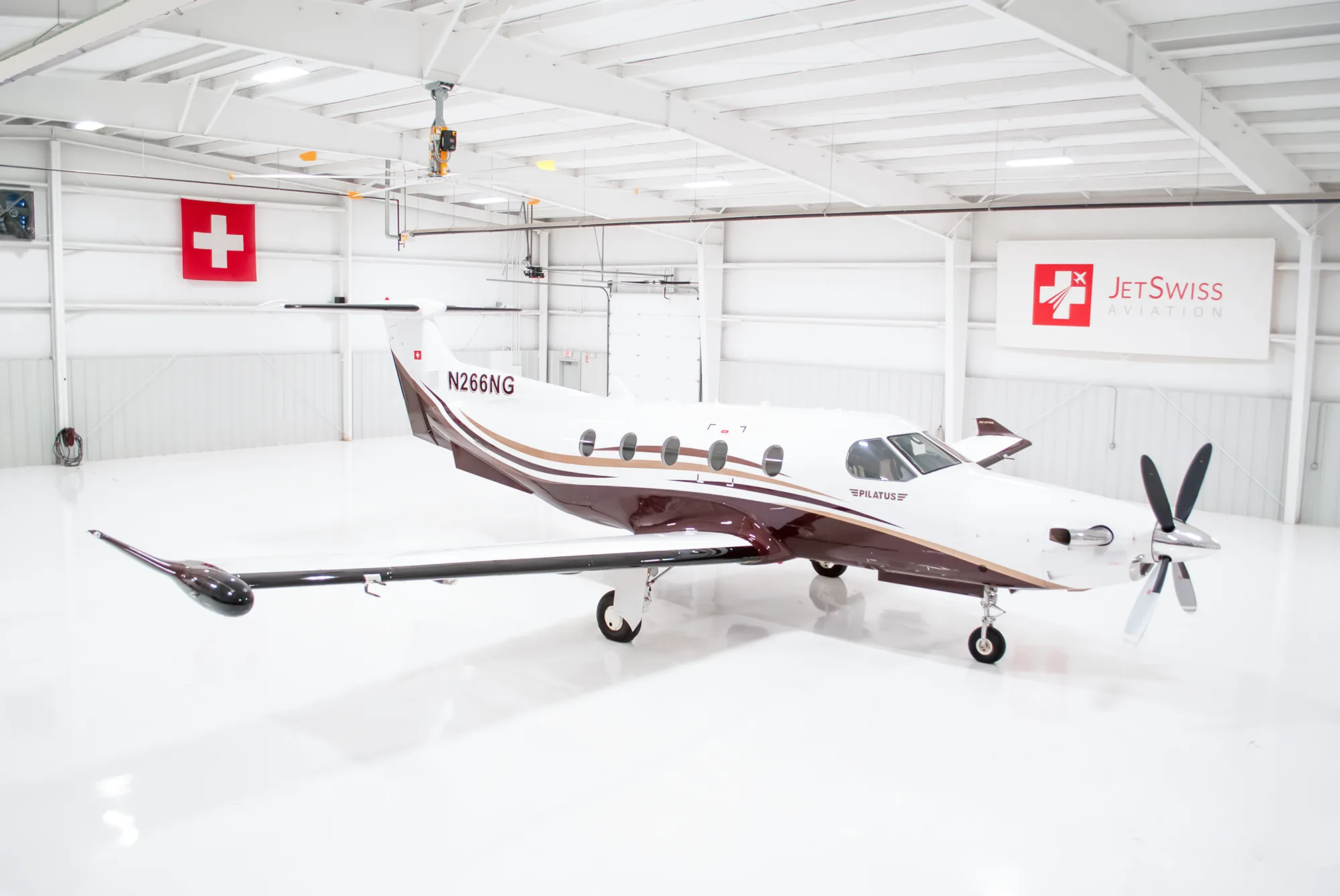 A Pilatus PC-12 NG aircraft with white and brown paint scheme, registration N266NG, parked in a brightly lit hangar.