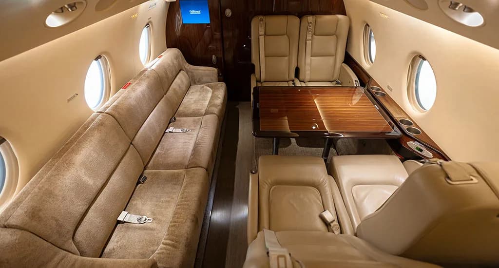 A view of the business jet's interior cabin, showing a long beige sofa, individual seats, and a polished wooden table.