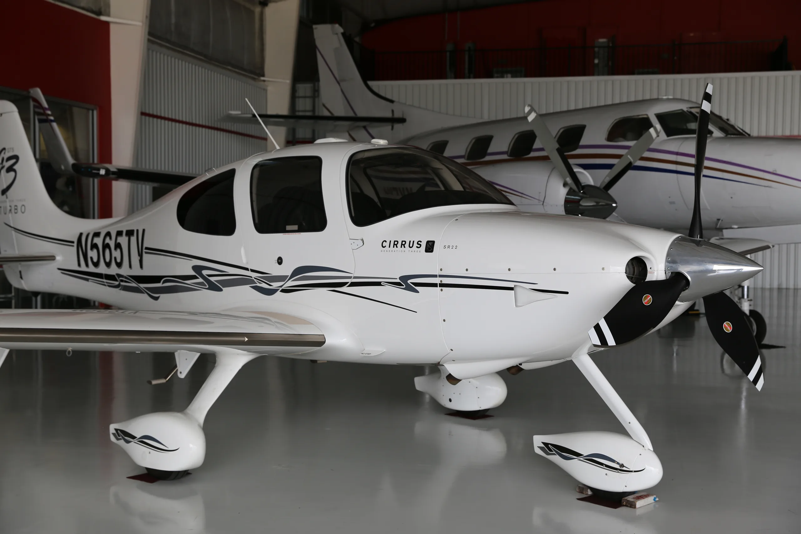 A white Cirrus SR22T aircraft, N565TV, parked inside a hangar next to another aircraft.