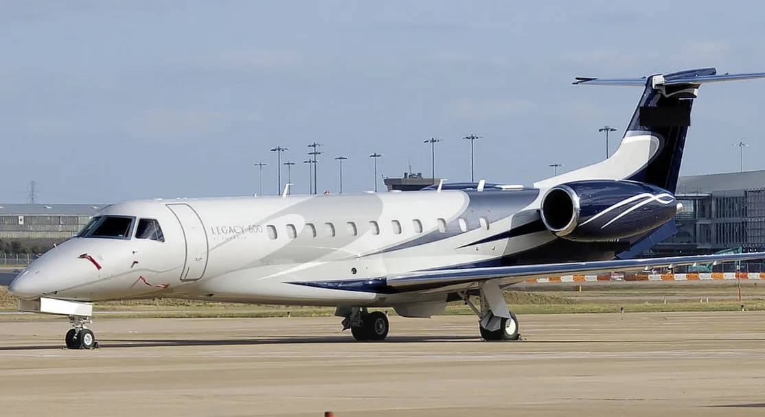 An Embraer Legacy 600 business jet with a distinctive grey, white, and dark blue paint scheme parked on an airport tarmac.