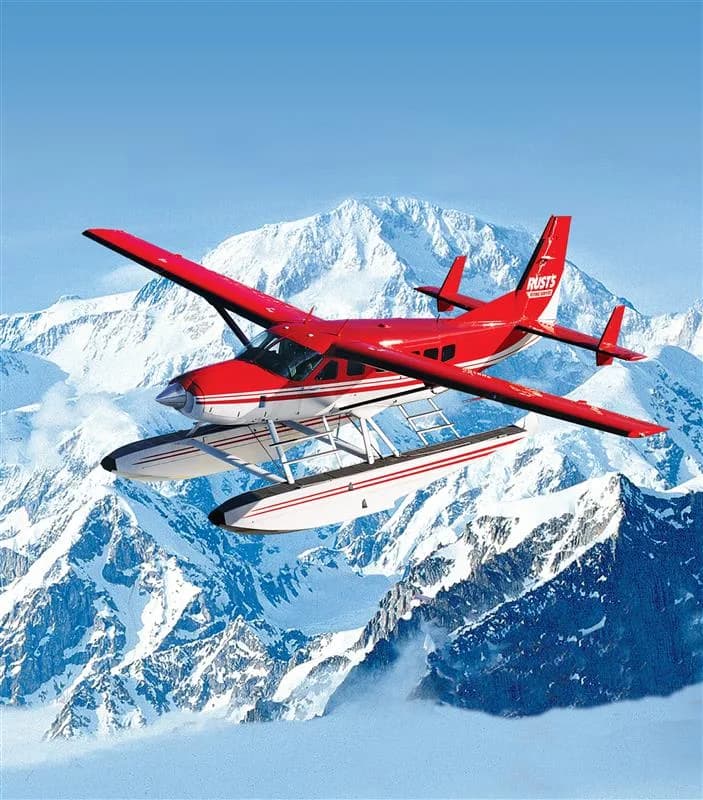 Red and white floatplane flying in front of a mountain.