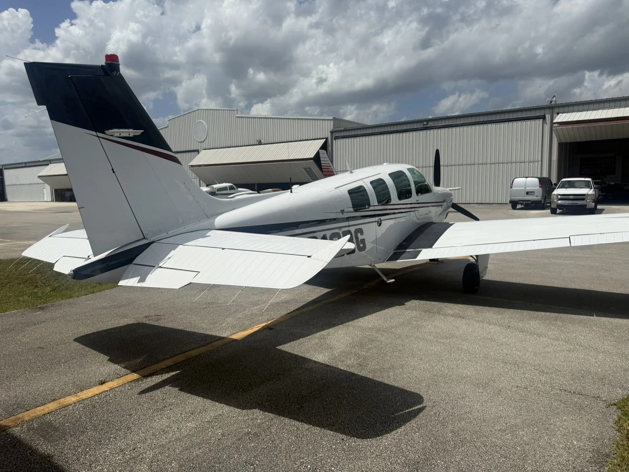 Rear view of a white Beechcraft Bonanza N812BG with dark red and black stripes parked on an airfield.