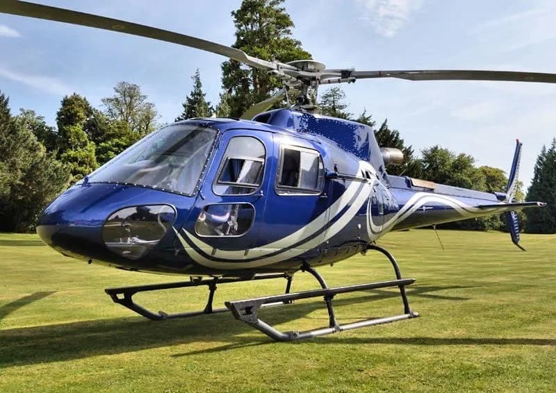 A blue helicopter with white and silver stripes is parked on a manicured green lawn with trees in the background.