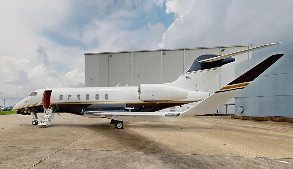 A white and black private jet with an open door and stairs on a tarmac next to a hangar under a cloudy sky.