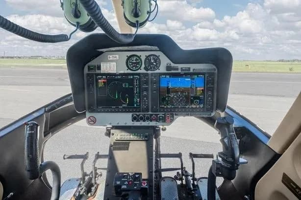 The cockpit of a helicopter showing a dual-screen glass panel display and flight controls.
