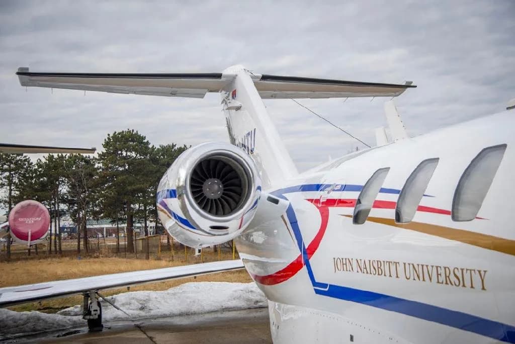 A close-up of the right engine and fuselage of a white business jet, showing "JOHN NAISBITT UNIVERSITY" text and a red, blue, and gold stripe.